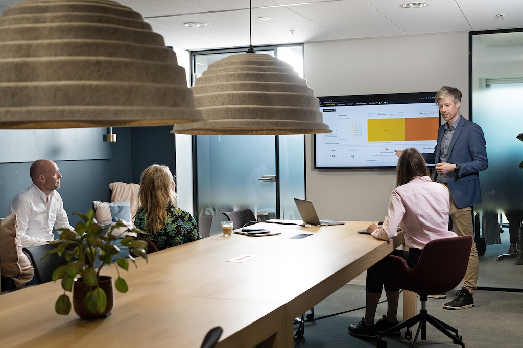 A group of four professionals is engaged in a meeting in a modern office space. A man in a blue blazer stands beside a large screen displaying graphs and data related to software composition analysis and open-source software, presenting to the team. Two women and another man sit attentively around a long wooden table with laptops and notebooks in front of them. The room is well-lit with natural light from the windows, and large, stylish pendant lights hang above the table. A potted plant adds a touch of greenery to the setting