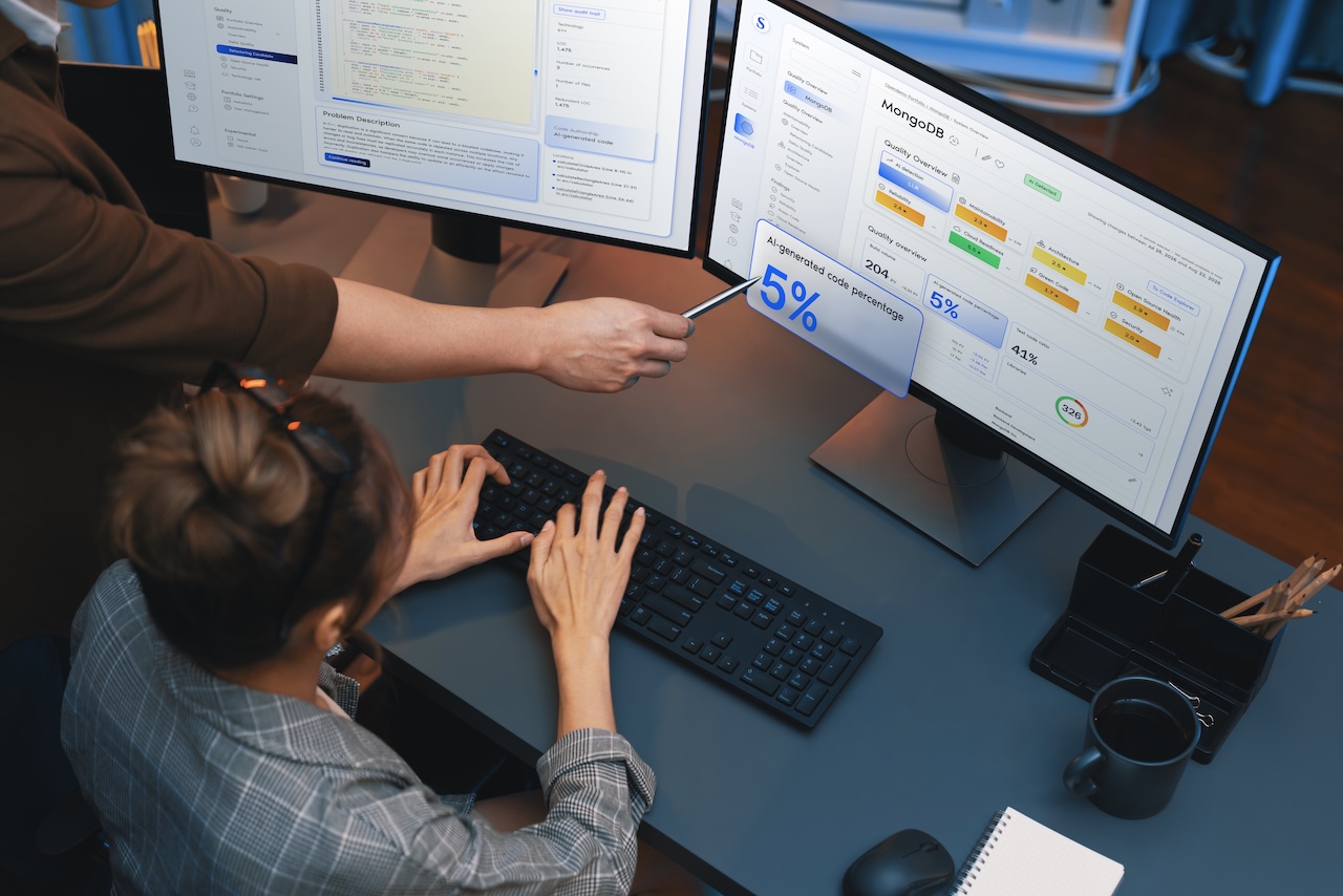 The image depicts a workspace environment featuring two large computer monitors on a desk. An individual, partially visible, points at the right monitor with a pen, indicating a section displaying statistics related to AI-generated code percentages. The screen shows various data and graphs with labels, such as "MongoDB" and "Quality Overview," suggesting a focus on data management. Another individual is seated at the desk, typing on a black keyboard. A notepad, a cup, and a pen holder with pencils are also visible on the desk, suggesting an active work setting. The overall color scheme is muted, with shades of blue and gray. Alt-text: Two monitors display coding and data statistics in a workspace. A person points at a screen with a pen while another types on a keyboard. Transcribed Text: MongoDB Quality Overview AI-generated code 5% 5% 41% 326