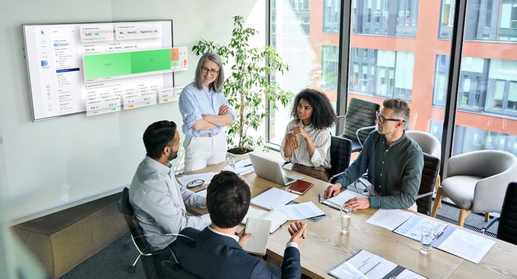 The image depicts a professional meeting in a modern conference room with large windows allowing natural light. Five individuals are gathered around a wooden table, engaged in discussion. A woman standing, dressed in a light blue shirt and white pants, appears to be leading the meeting. The seated participants include two men and one woman on one side, and another man on the other side, all taking notes or using laptops. On the wall, a large screen displays a digital interface with charts and code, suggesting the meeting is about an AI-assisted coding platform. The room includes a potted plant and comfortable chairs, with a view of an office building through the windows. Alt-text: A meeting in a modern conference room with five people discussing in front of a screen displaying AI-assisted coding solutions. Transcribed Text: Partial text on the digital interface includes "AI-generated code" and "ML, GenAI, Agentic AI." Additional text on the interface and documents is not clearly visible.