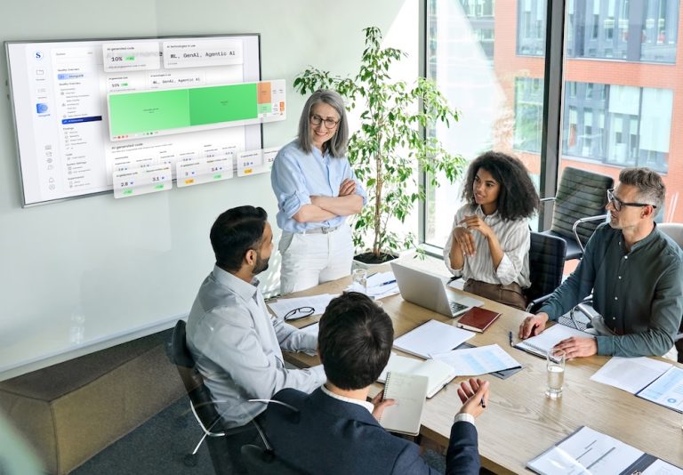 The image depicts a professional meeting in a modern conference room with large windows allowing natural light. Five individuals are gathered around a wooden table, engaged in discussion. A woman standing, dressed in a light blue shirt and white pants, appears to be leading the meeting. The seated participants include two men and one woman on one side, and another man on the other side, all taking notes or using laptops. On the wall, a large screen displays a digital interface with charts and code, suggesting the meeting is about an AI-assisted coding platform. The room includes a potted plant and comfortable chairs, with a view of an office building through the windows. Alt-text: A meeting in a modern conference room with five people discussing in front of a screen displaying AI-assisted coding solutions. Transcribed Text: Partial text on the digital interface includes "AI-generated code" and "ML, GenAI, Agentic AI." Additional text on the interface and documents is not clearly visible.
