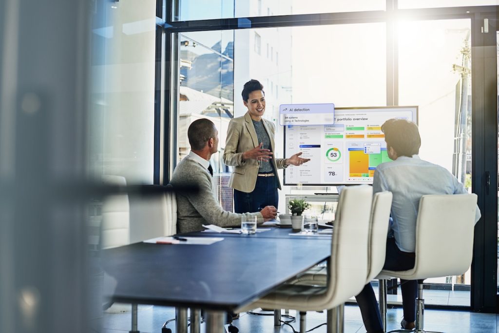 The image depicts an office meeting scene with three individuals in a modern glass-walled conference room. A woman in business attire is standing and presenting information displayed on a large screen, which shows various colorful graphs and data metrics related to AI detection and portfolio overview. Two men are seated at a conference table listening attentively, with notebooks, a laptop, and a small plant on the table. The room is well-lit with natural light streaming in through large windows, creating a bright, open atmosphere. Outside, buildings and clear skies are visible, adding a sense of connectivity to the urban environment. Alt-text: An office meeting with a woman presenting data on a screen while two men listen. Transcribed Text: AI detection 34 systems Using AI Technologies portfolio overview
