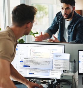 The image depicts two men in a discussion in a modern office setting, centered around a computer screen displaying an AI-assisted coding platform. The man in the foreground is wearing a beige shirt and glasses, his back turned to the camera. The man across from him is gesturing with his hands, wearing a denim jacket over a white T-shirt. The computer monitor shows a coding interface with highlighted sections and text boxes. The workspace is bright, with natural light coming through large windows, plants, and office equipment visible in the background. An interface on the screen includes various coding options, file directories, and comments, emphasizing collaboration and problem-solving. Alt-text: Two men discussing in front of a computer displaying an AI-assisted coding platform. Transcribed Text: Problem Description "In src, duplication is a significant concern because it can lead to a bloated codebase, making it challenging to read and maintain. When the same code is repeated across multiple locations, the risk of having inconsistent results is significantly increased. This inconsistency makes coordinating future releases, as well as performing security audits, more tedious. The process of identifying replication allows the engineering team to focus on areas of the code that may require attention. Duplications can also hinder the ability to update or on efficiency as the effort required is wasted on issues aimed not to be cultivated."
