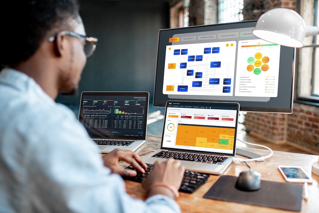 The image shows a person sitting at a desk using two laptops and a large monitor. The individual is focused on the screens in front of them, which display various analytics and charts from the Sigrid software platform. The large monitor features a software architecture diagram, displaying a flowchart with boxes and connecting lines, alongside a circular diagram with various colored sections. Both laptops have screens filled with data and graphs. One laptop screen consists of bar and line graphs with a dark background, while the other shows a dashboard interface with orange, red, and green sections. The setting includes a brick wall in the background and a large lamp on the desk. There is a smartphone and a mouse next to the laptops.