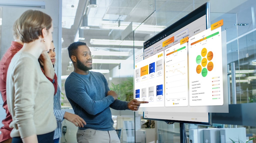 This is a mockup of group of four people in an office discussing a software architecture & code quality on a large digital screen displaying colorful graphs and charts.