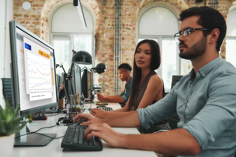 This is a mockup image of three people working at computers in a brick-walled office with charts displayed on screens.