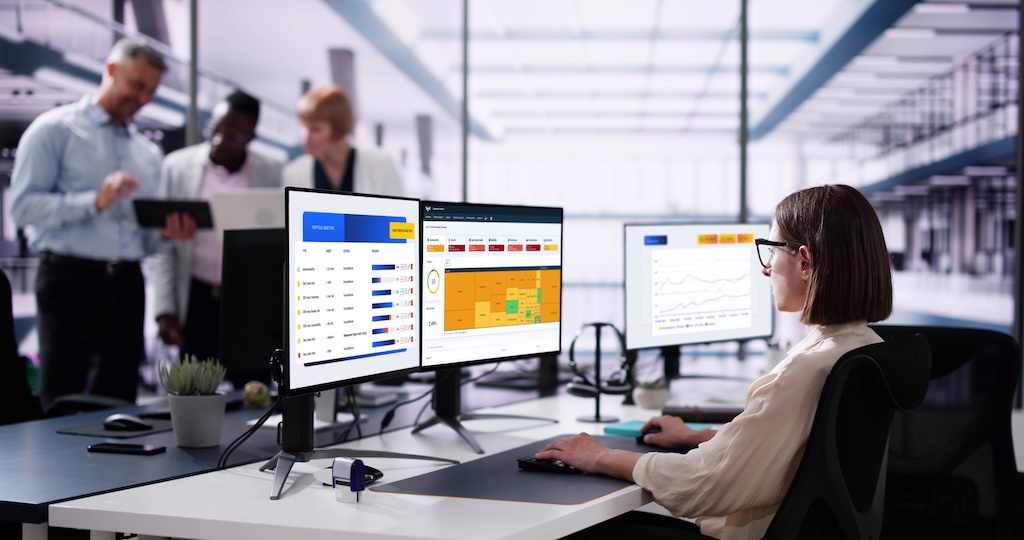 This is a mockup image of woman working at a desk with two monitors displaying the Sigrid's feature portfolio objectives' charts and graphs in an open-plan office .