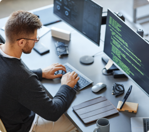 Man typing on a laptop with dual monitors displaying code.