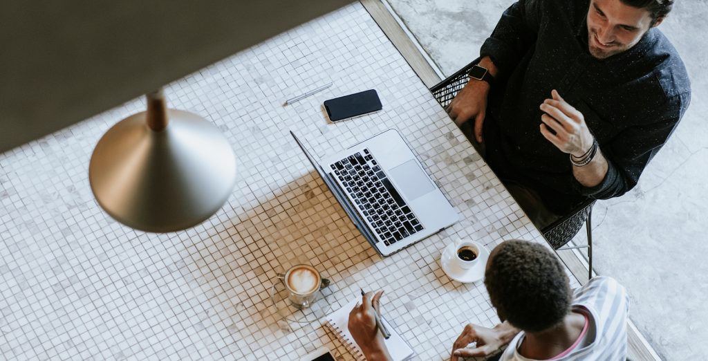 Overhead view of two people at a tiled table with a laptop, phone, notebook, and coffee.