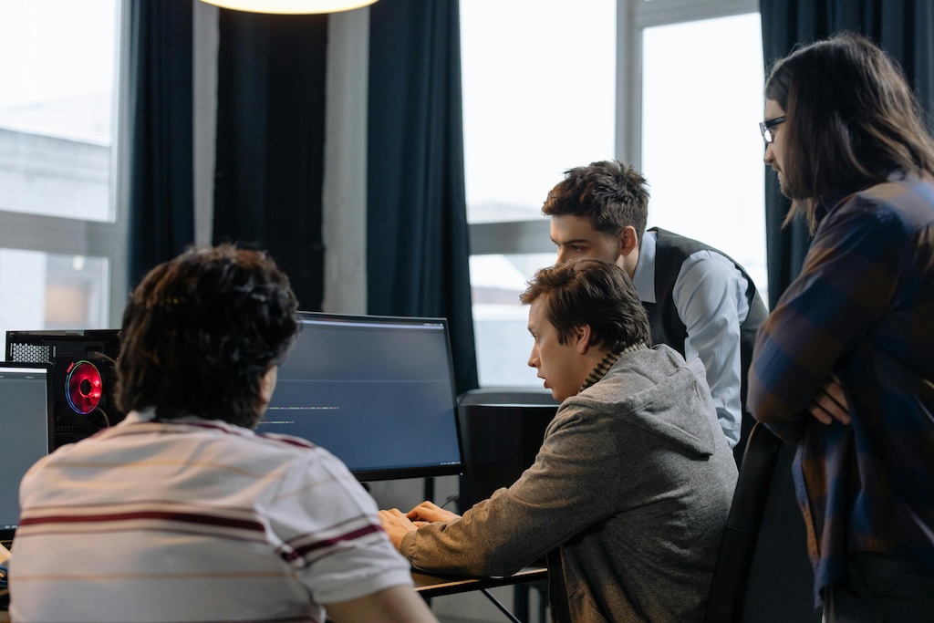 A group of four people collaborating in front of a computer in a modern office setting. One person is seated, typing on the keyboard, while the other three stand closely behind, looking at the monitor. The scene suggests teamwork and focused problem-solving, likely involving programming or technical work. The office has large windows and a well-lit interior, contributing to a professional and collaborative atmosphere.