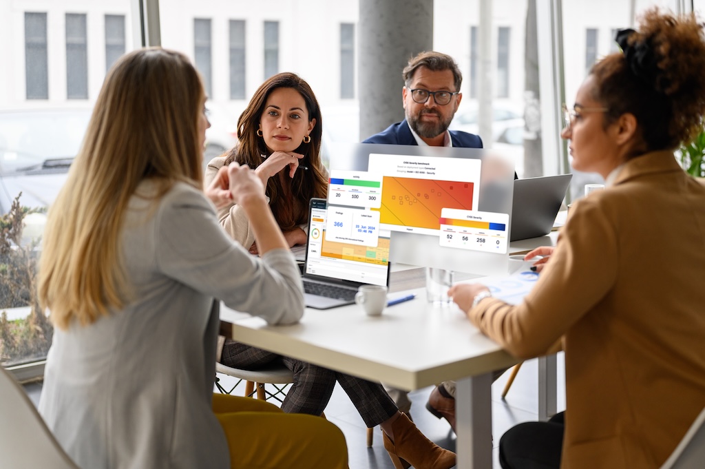 This is a mockup of four people having a meeting in an office with laptops displaying graphs & data in Sigrid security feature.