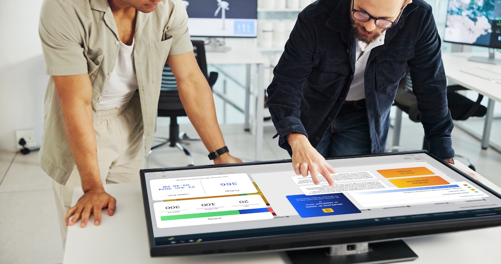 This is a mockup image of two people interacting with a large touchscreen monitor displaying colorful interface elements in an office.