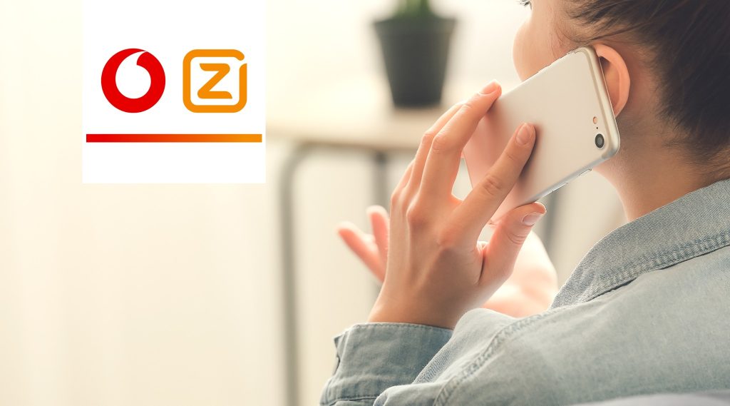 A woman holding a smartphone to her ear while on a call, with the Vodafone Ziggo logo displayed in the top left corner. The background is softly lit with a neutral color, and a plant is visible in the distance.