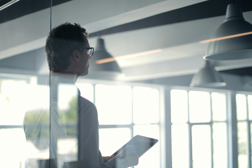 We see an executive through the window in an office setting looking at a detailed report on his Ipad.