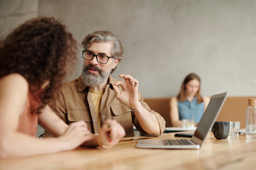 he image shows two individuals sitting at a wooden table, engaged in conversation. The person on the right, with a gray beard and glasses, gestures with his right hand, suggesting active discussion. He is wearing a brown jacket over a mustard-colored shirt. The person on the left has curly hair and is attentively listening, slightly leaning in. On the table is an open laptop, a black mug, and a clear glass with some water. In the background, another individual can be seen blurred, working on a laptop, giving the setting a casual and collaborative feel.