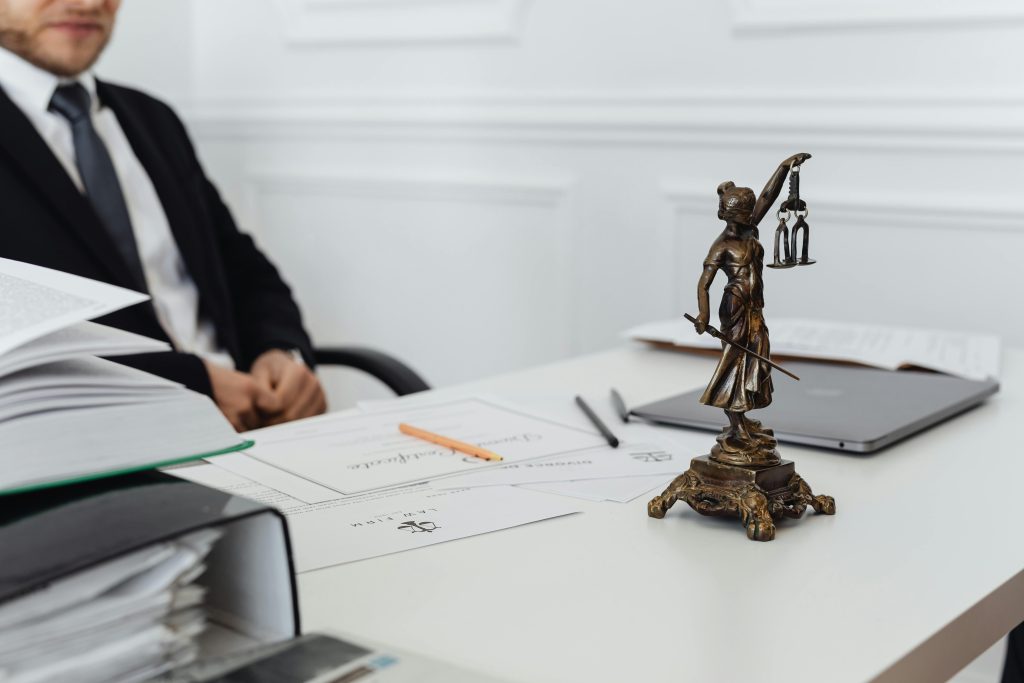 A bronze Lady Justice statue on a desk with office items and a person seated in the background.