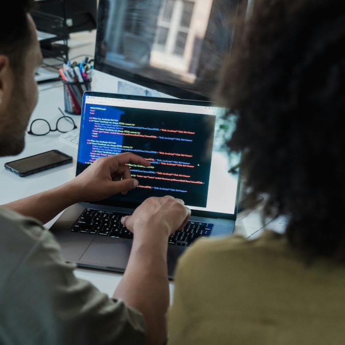 This is a mockup image of two people looking at code on a laptop in an office setting.