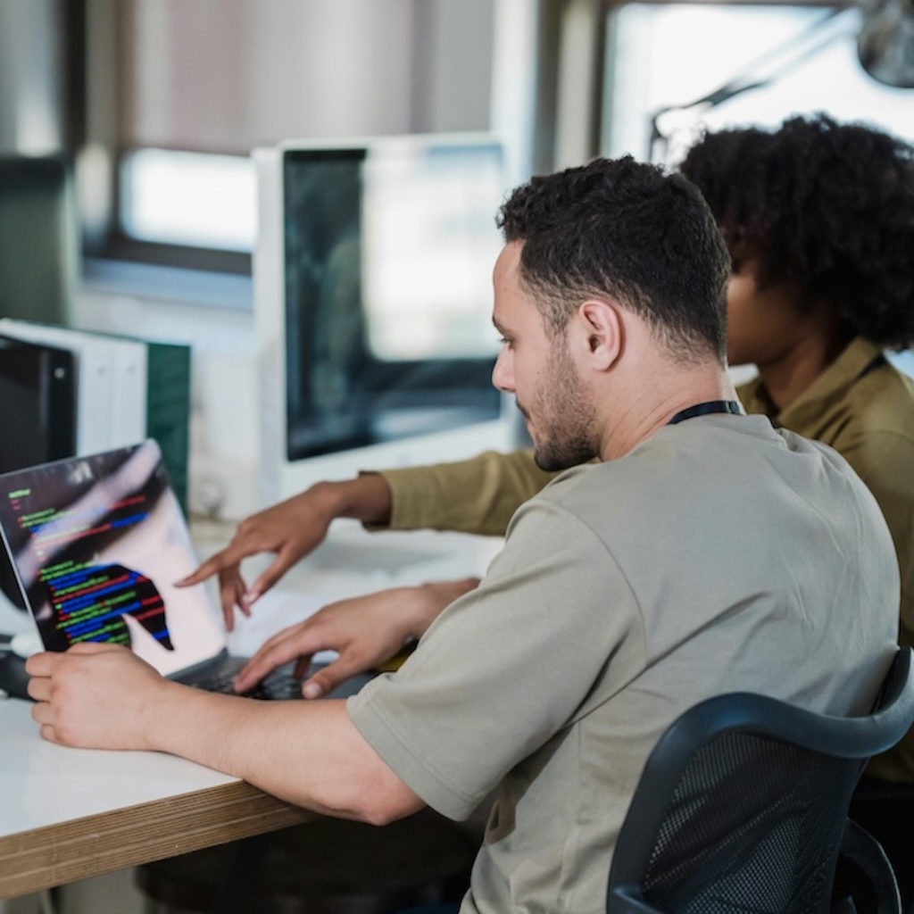 This is a mockup image of two people collaborating on a coding project at a desk with a laptop displaying code.