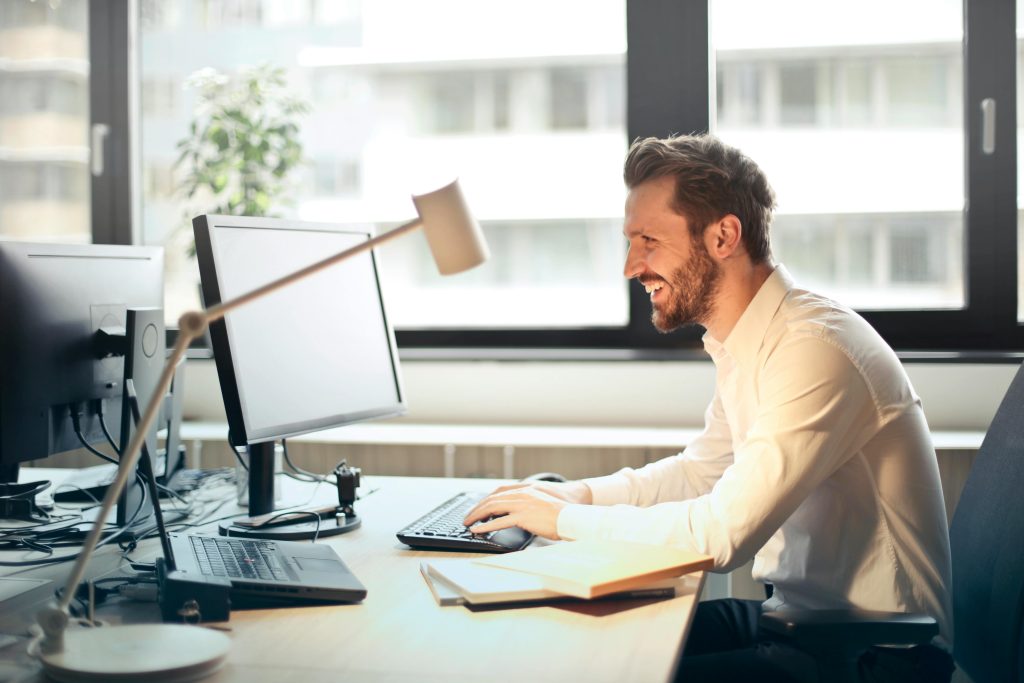 Image of a man smiling while working at a computer in a bright office with two monitors and a laptop.