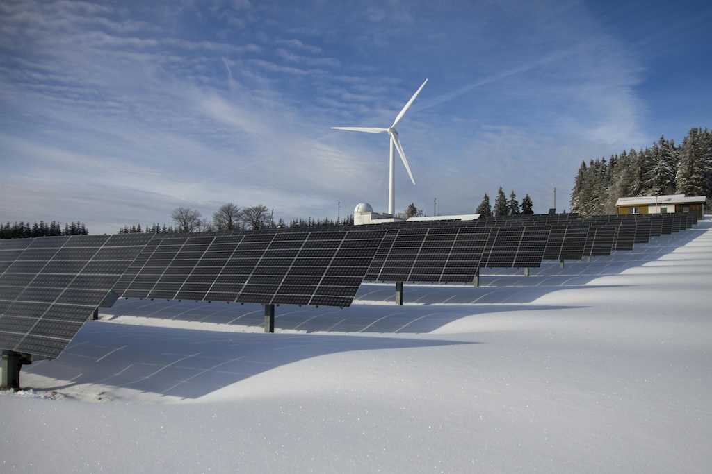 Solar panels in a snowy field with a wind turbine and evergreen trees in the background.