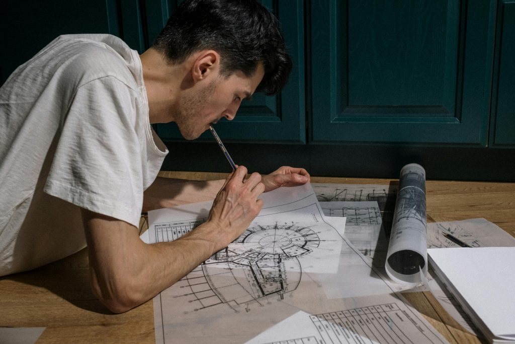 A man leans over a wooden table, pencil in hand, studying detailed architectural blueprints spread across the surface. A rolled drawing sits nearby.