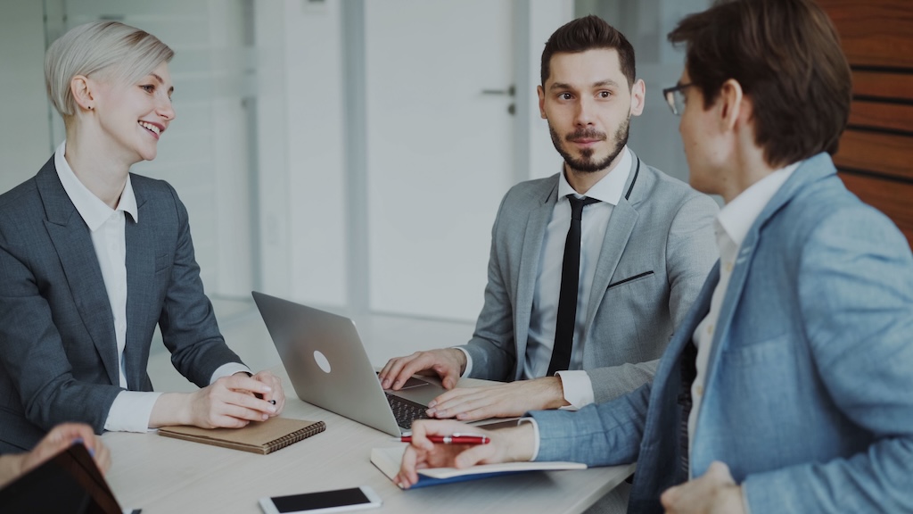 The image depicts a business meeting taking place in a bright and modern office environment. Three individuals are seated around a light wooden table. On the left, a woman with short, platinum blonde hair and a confident smile is dressed in a dark gray suit with a white blouse. She is engaging with the others while holding her hands in front of her. In the center, a man in a light gray suit with a black tie and neatly groomed beard is focused on a laptop in front of him, looking up with a thoughtful expression. On the right, another man in a light blue suit is turned towards the center man, seemingly explaining something while holding a red pen in his right hand. The setting has a neutral and minimalistic aesthetic, emphasizing the professionalism of the situation. The background features transparent glass, allowing natural light to illuminate the scene.