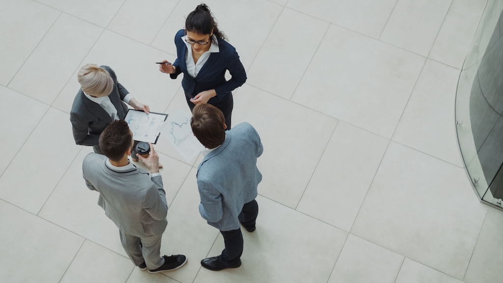 The image captures a top-down view of four business professionals engaged in a discussion. They are standing in a spacious, well-lit area with light-colored tiled flooring. The individuals are dressed in formal business attire. The woman in the center, wearing a dark suit, is holding a pen and pointing towards a document spread on a flat surface in front of her, suggesting she is leading the conversation. Two men, one in a light gray suit and the other in a blue suit, are looking towards the document. The man in the light gray suit holds a coffee cup in one hand. The fourth person, a woman with blonde hair, is wearing a white blouse under a gray jacket and is also observing the document. The overall atmosphere conveys a collaborative and professional setting, with a focus on teamwork.