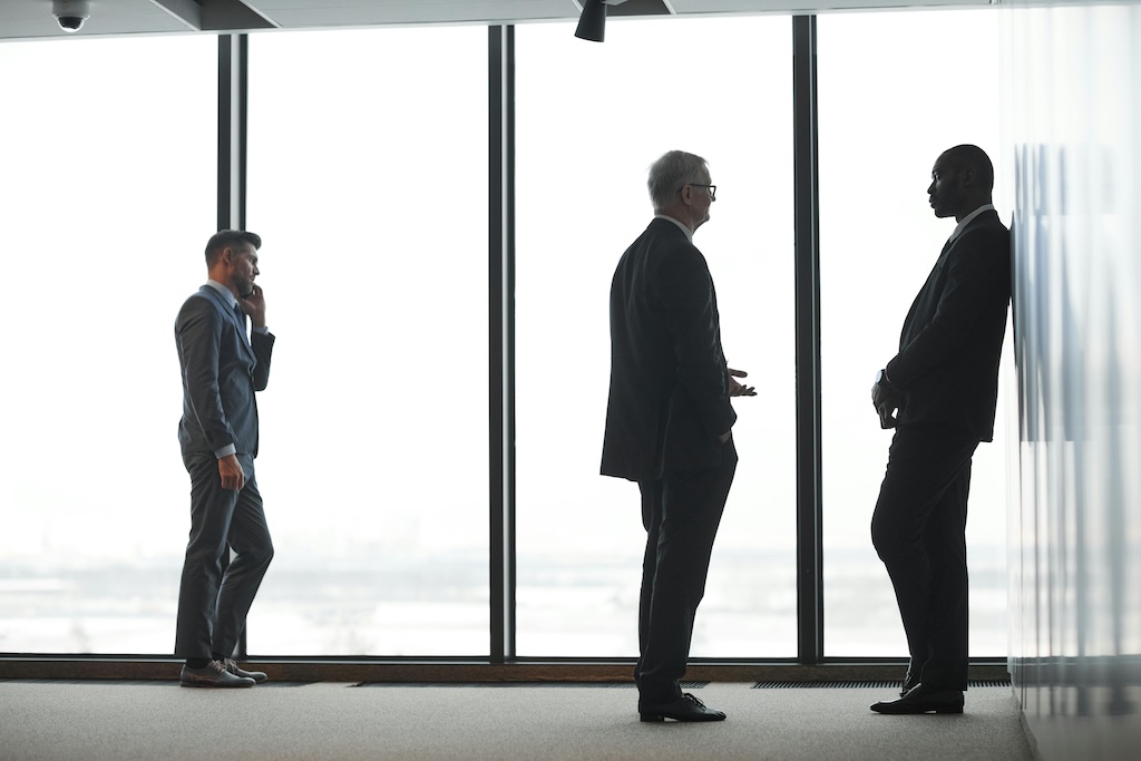 A corporate hallway with large windows. One person is on the phone, two colleagues are discussing important matters.