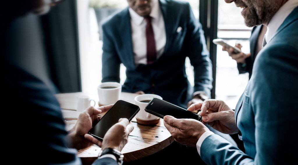 Men in suits holding smartphones around a table with coffee cups having a discussion.