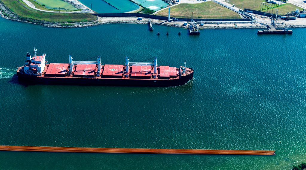 Aerial view of a red cargo ship in a waterway with a shoreline and port facilities in the background.